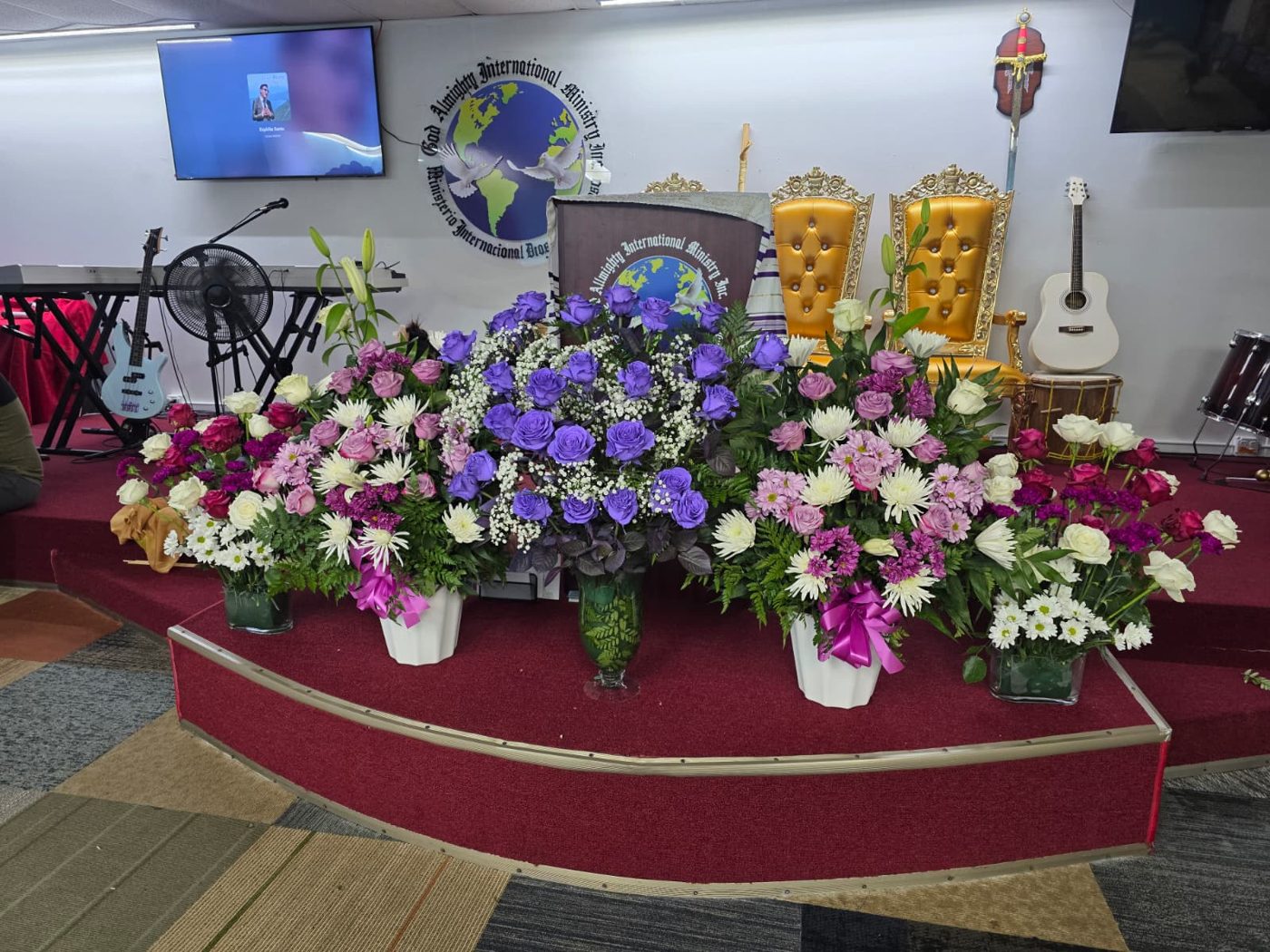 Altar de la iglesia adornado con arreglos de rosas moradas, blancas y rosadas frente al logo del ministerio